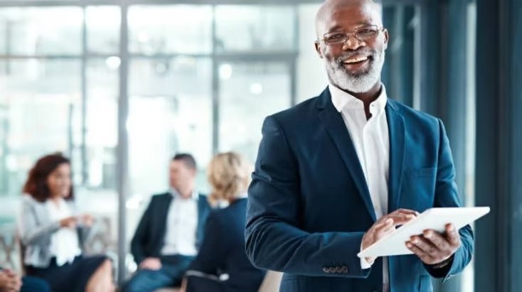 A smiling business advisor holding a tablet, with a team meeting behind him
