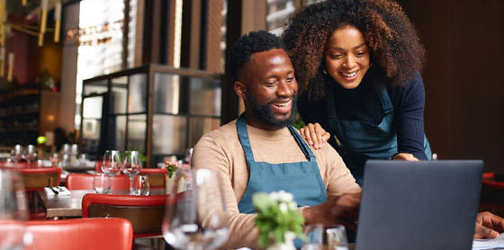 Two smiling business owners in aprons looking at a laptop in their restaurant
